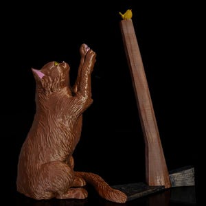 May include: A brown cat figurine sitting on its haunches with front paws raised toward a small yellow bird perched atop a tall, thin, brown wooden post. The cat has pink inner ears and textured fur, while the post stands on a dark base.