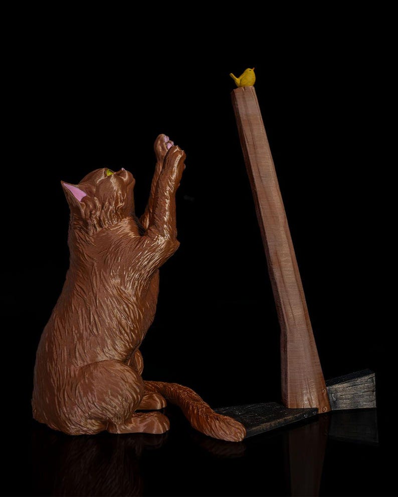 May include: A brown cat figurine sitting on its haunches with front paws raised toward a small yellow bird perched atop a tall, thin, brown wooden post. The cat has pink inner ears and textured fur, while the post stands on a dark base.