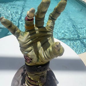 May include: A sculpted, upright zombie hand figurine featuring textured, pale green skin with visible wrinkles, a severed finger with a gold ring, a dark wristband, and a red, exposed wound detail, all mounted on a circular, dark-colored base.