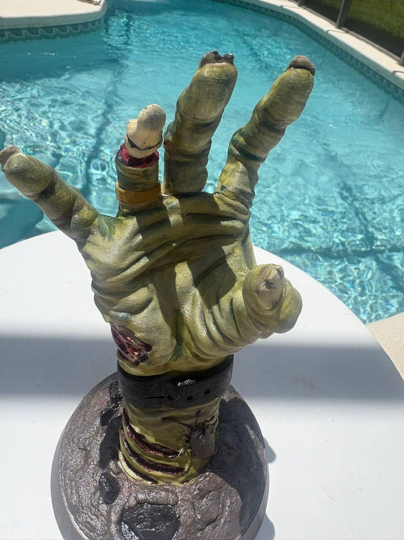 May include: A sculpted, upright zombie hand figurine featuring textured, pale green skin with visible wrinkles, a severed finger with a gold ring, a dark wristband, and a red, exposed wound detail, all mounted on a circular, dark-colored base.