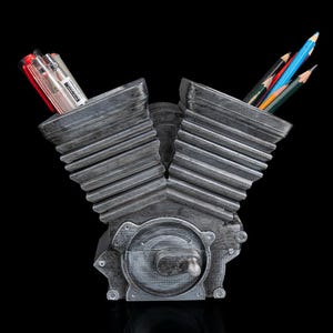 May include: A gray V-twin engine shaped desk organizer with a metallic finish. The two cylinder heads serve as containers for pens and pencils. The base features a circular engine cover detail with a small protruding lever, all set against a black background.