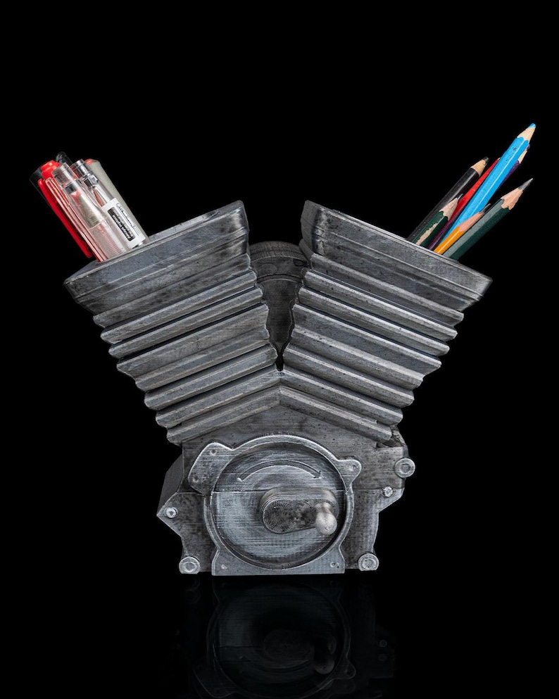 May include: A gray V-twin engine shaped desk organizer with a metallic finish. The two cylinder heads serve as containers for pens and pencils. The base features a circular engine cover detail with a small protruding lever, all set against a black background.