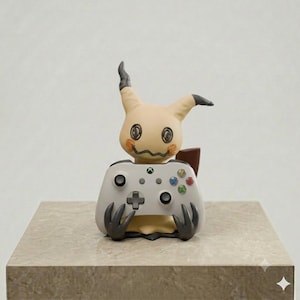Op de afbeelding: Een beige Mimikyu-figuurtje met donkergrijze oortoppen en gekrabbelde gelaatstrekken, dat een lichtgrijze videogamecontroller vasthoudt. De controller is voorzien van een richtingspad, twee analoge sticks, vier gekleurde actieknoppen en een centraal Xbox-logo.