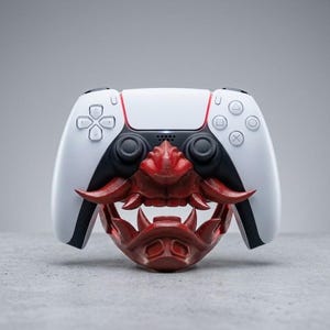 May include: A white and black video game controller resting on a dark red, sculpted mask stand. The stand features a detailed design with prominent fangs, a nose bridge, and curved tusks that cradle the controller in a central position.