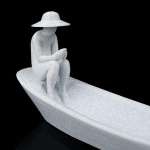 May include: A light gray, speckled, 3D-printed figurine of a person wearing a wide-brimmed hat, sitting inside a small boat. The figure is positioned at the bow of the vessel, holding a small object in their hands while looking downward.