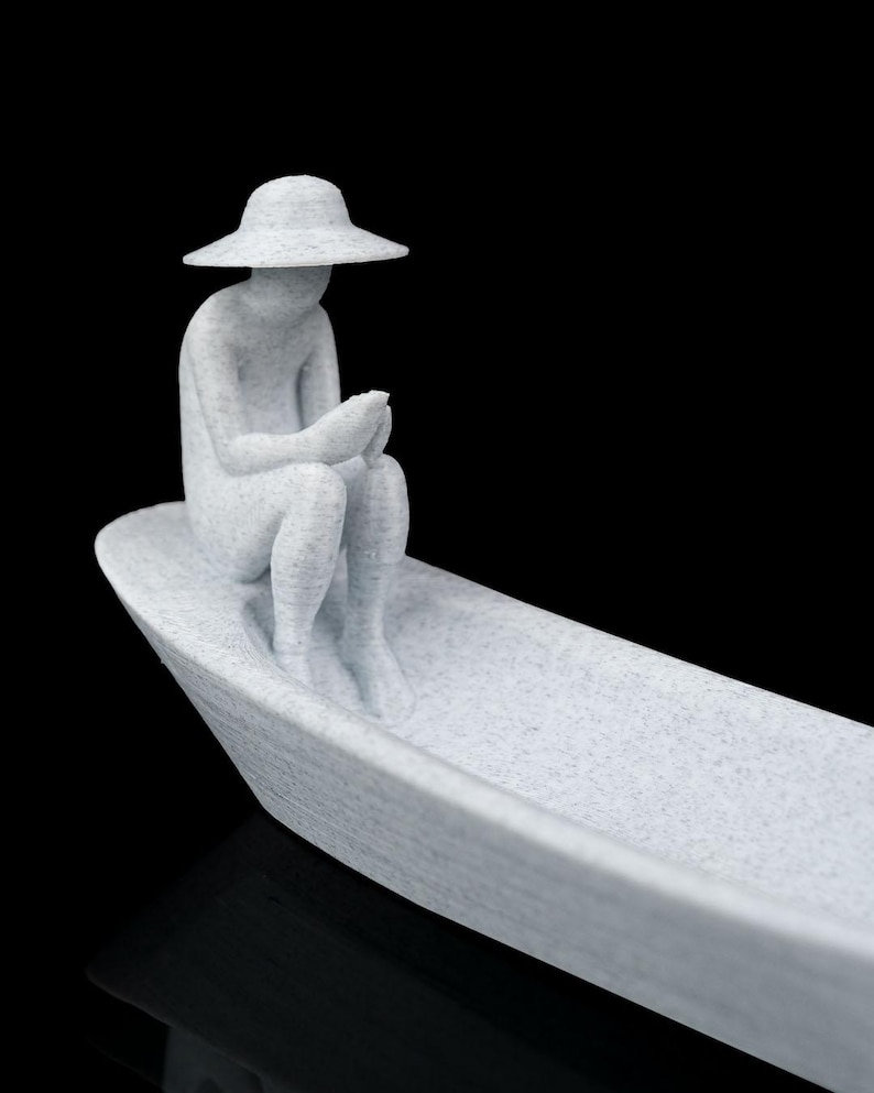 May include: A light gray, speckled, 3D-printed figurine of a person wearing a wide-brimmed hat, sitting inside a small boat. The figure is positioned at the bow of the vessel, holding a small object in their hands while looking downward.