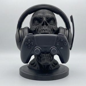 May include: A matte black skull-shaped stand designed to hold gaming accessories. The skull features a headset resting on its cranium and a black video game controller positioned in its mouth, all mounted on a circular base.