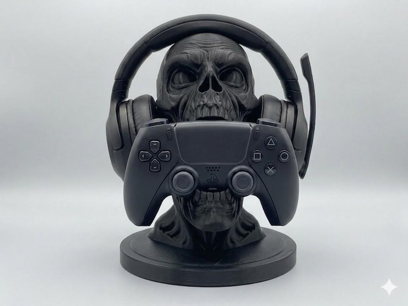 May include: A matte black skull-shaped stand designed to hold gaming accessories. The skull features a headset resting on its cranium and a black video game controller positioned in its mouth, all mounted on a circular base.