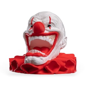 May include: A white clown head sculpture with a large red nose, red vertical lines above the eyes, and a wide open mouth revealing white teeth. The figure is set upon a ruffled red collar base, featuring a textured surface and bold colours.