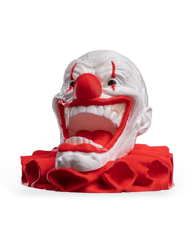 May include: A white clown head sculpture with a large red nose, red vertical lines above the eyes, and a wide open mouth revealing white teeth. The figure is set upon a ruffled red collar base, featuring a textured surface and bold colours.