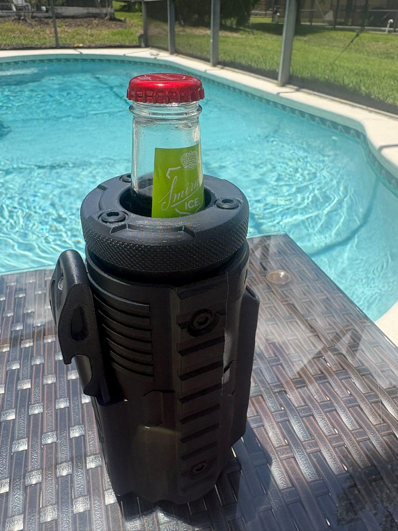 Pode incluir: Um refrigerador de bebidas cil&iacute;ndrico preto com um exterior texturizado de estilo t&aacute;tico, apresentando trilhos verticais e um abridor de garrafas lateral. Uma garrafa de vidro com tampa vermelha e r&oacute;tulo verde Smirnoff ICE est&aacute; inserida.