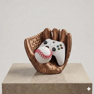 Puede incluir: Una escultura de guante de b&eacute;isbol color bronce que sostiene una pelota de b&eacute;isbol blanca con costuras rojas y un mando de videojuego blanco con joysticks negros y botones de acci&oacute;n multicolores. El guante tiene un acabado texturizado.