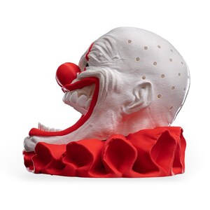 May include: A white clown head sculpture with a bright red nose, wide mouth showing white teeth, and a red ruffled collar. The bald head features a textured surface with small, evenly spaced circular indentations across the top and sides.