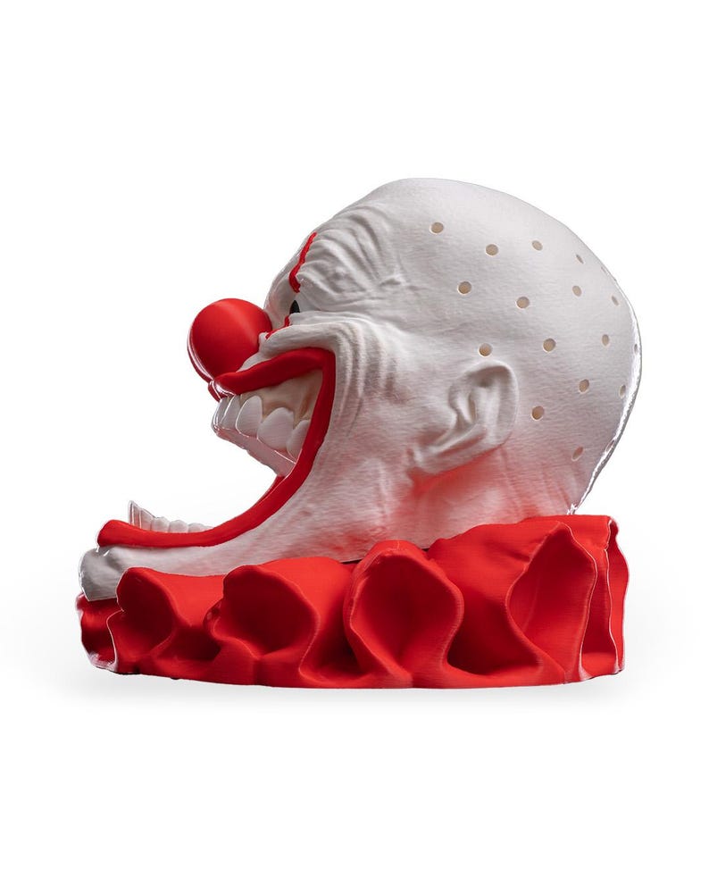 May include: A white clown head sculpture with a bright red nose, wide mouth showing white teeth, and a red ruffled collar. The bald head features a textured surface with small, evenly spaced circular indentations across the top and sides.