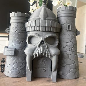 Op de afbeelding: Een grijze 3D-geprinte kasteelstructuur met een centraal poortgebouw in de vorm van een schedel, geflankeerd door twee torens met een steenstructuur. De schedel heeft grote oogkassen en scherpe hoektanden rondom een deur met een doodskop.