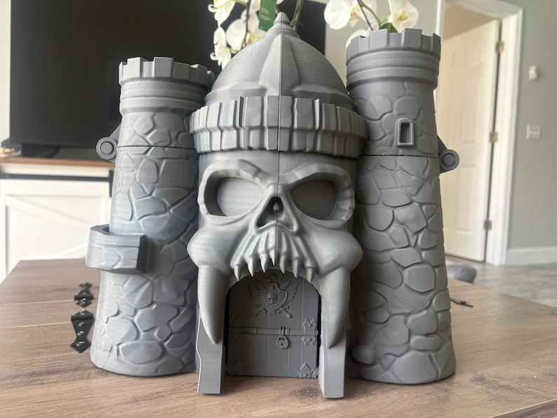 Op de afbeelding: Een grijze 3D-geprinte kasteelstructuur met een centraal poortgebouw in de vorm van een schedel, geflankeerd door twee torens met een steenstructuur. De schedel heeft grote oogkassen en scherpe hoektanden rondom een deur met een doodskop.