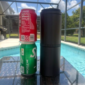 以下が含まれることがあります： マットブラックの背の高い円筒形キャニスターが、2つのアルミ製飲料缶の隣に置かれています。上の缶は赤色で白いコカ・コーラのロゴと栄養成分表示があり、下の缶は緑色で白いスプライトのロゴとリサイクルに関するテキストが記載されています。