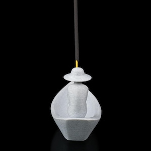 May include: A light gray 3D-printed incense holder shaped like a small boat with a figure wearing a wide-brimmed hat seated inside. A thin yellow stick of incense is inserted into the top of the hat, extending vertically upward against a black background.