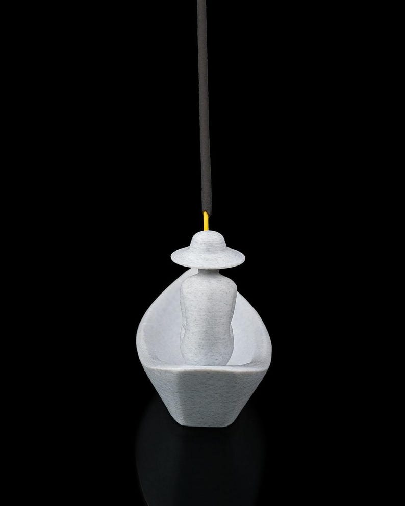 May include: A light gray 3D-printed incense holder shaped like a small boat with a figure wearing a wide-brimmed hat seated inside. A thin yellow stick of incense is inserted into the top of the hat, extending vertically upward against a black background.
