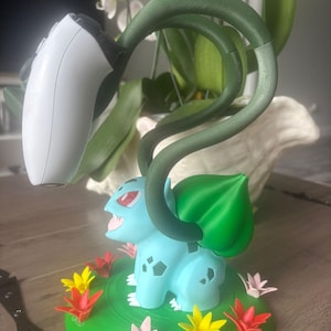 Op de afbeelding: Een lichtblauw Bulbasaur-figuur op een ronde groene basis versierd met kleine rode, gele en roze bloemen. Het figuur heeft twee lange, donkergroene ranken die omhoog reiken om een wit en zwart videospel-controller vast te houden.