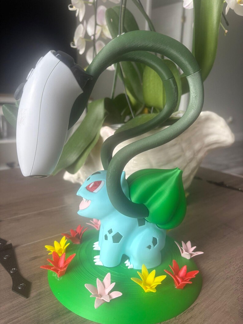 Op de afbeelding: Een lichtblauw Bulbasaur-figuur op een ronde groene basis versierd met kleine rode, gele en roze bloemen. Het figuur heeft twee lange, donkergroene ranken die omhoog reiken om een wit en zwart videospel-controller vast te houden.