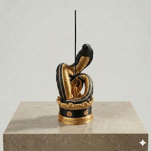 May include: A black and gold incense holder featuring a coiled snake sculpture atop a circular base. The snake has a textured black exterior and a smooth gold underside. A thin, black incense stick stands vertically in the centre holder.