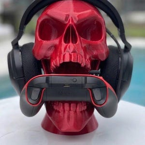 May include: A glossy red 3D-printed human skull model serving as a stand, featuring black over-ear headphones resting on the cranium and a black and red wireless gaming controller held firmly within the open jaw of the skull structure.