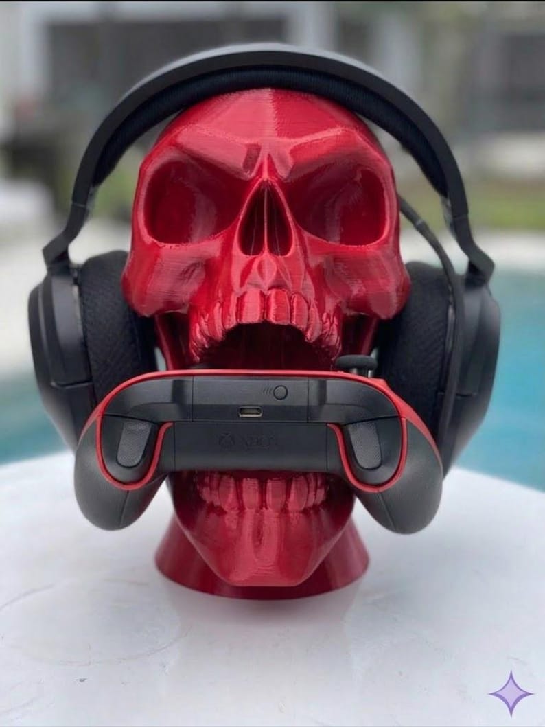 May include: A glossy red 3D-printed human skull model serving as a stand, featuring black over-ear headphones resting on the cranium and a black and red wireless gaming controller held firmly within the open jaw of the skull structure.