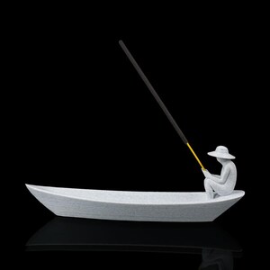 May include: A light gray textured incense holder shaped like a small boat with a seated figure wearing a wide-brimmed hat. The figure holds a long, thin yellow and black incense stick that extends upward from the boat, creating a fishing rod effect.