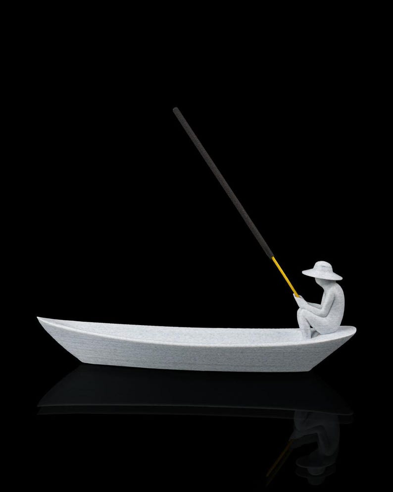 May include: A light gray textured incense holder shaped like a small boat with a seated figure wearing a wide-brimmed hat. The figure holds a long, thin yellow and black incense stick that extends upward from the boat, creating a fishing rod effect.