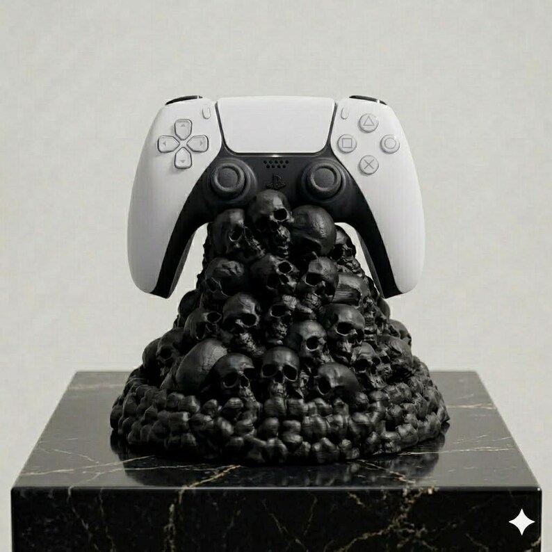 Op de afbeelding: Een wit en zwart videogame-controller rustend op een gebeeldhouwde basis bestaande uit een dichte stapel kleine, matzwarte schedels. De controller is voorzien van standaard richtingsknoppen, analoge sticks en actiesymbolen op de voorzijde.