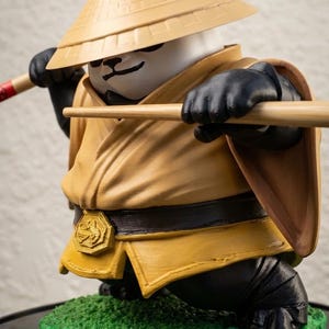 May include: A panda figurine wearing a tan conical hat and a tan robe with a dark brown belt featuring a gold hexagonal emblem. The panda holds a long wooden staff in its black hands and stands on a textured green base with a black circular edge.