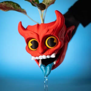 May include: A bright red devil-shaped planter with large yellow eyes, white teeth, and a blue tongue extending downwards. The planter features two horns on top and holds a small green plant with variegated leaves, set against a solid blue background.