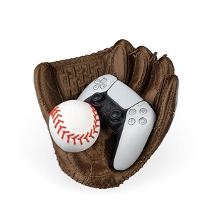 Puede incluir: Un guante de b&eacute;isbol marr&oacute;n texturizado que sostiene una pelota de b&eacute;isbol blanca con costuras rojas y un mando de videojuego inal&aacute;mbrico blanco y negro. El mando cuenta con una cruceta, dos joysticks anal&oacute;gicos y cuatro botones de acci&oacute;n.