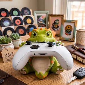 May include: A green dragon-shaped controller stand holding a white wireless gaming controller. The dragon has orange spots, a cream-colored belly, and large eyes. The stand is designed to securely cradle the controller in its mouth and arms.
