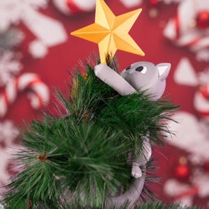 May include: A grey cat figurine climbing a green artificial pine tree and reaching up to place a bright yellow five-pointed star on the top. The cat has white inner ears and large black eyes, with its body partially obscured by the pine needles.