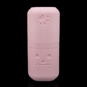May include: A cylindrical light pink container with a smooth matte finish. The top section features an embossed paw print design, while the bottom section displays an embossed cat face with two circular eyes, a small nose, and a mouth.
