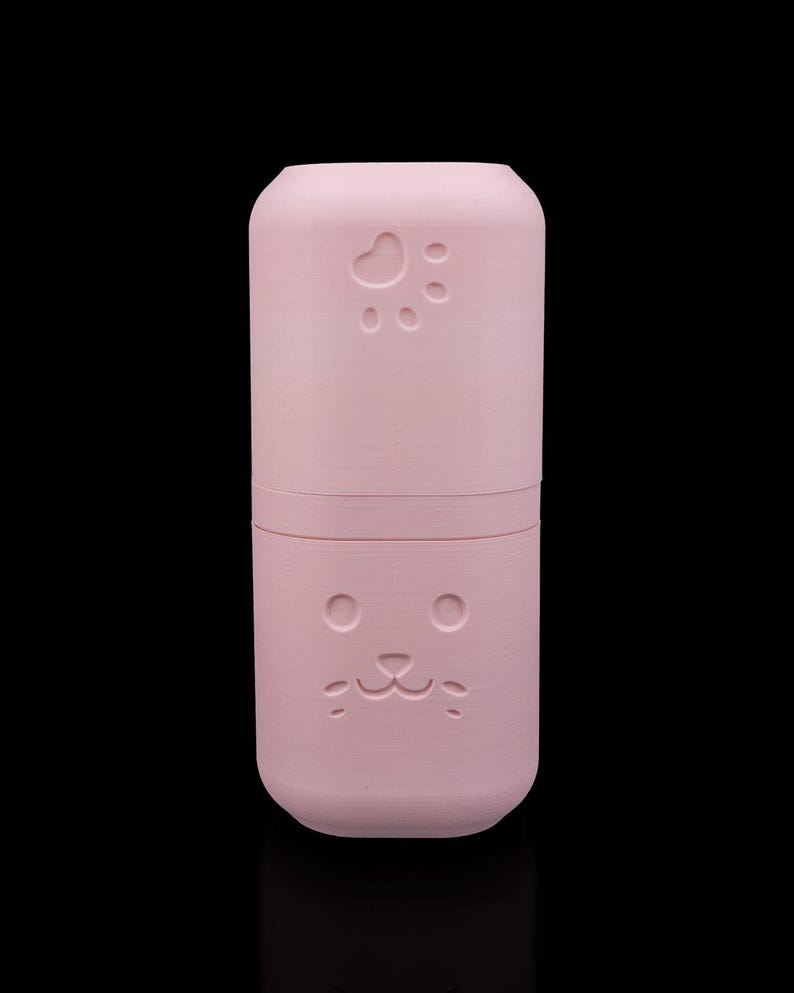 May include: A cylindrical light pink container with a smooth matte finish. The top section features an embossed paw print design, while the bottom section displays an embossed cat face with two circular eyes, a small nose, and a mouth.