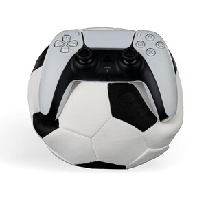 Op de afbeelding: Een wit en zwart voetbal pluche speelgoed met een witte en zwarte draadloze videogamecontroller die er bovenop rust. De controller is voorzien van een centraal touchpad, twee analoge sticks, een D-pad en vier actieknoppen met symbolen.