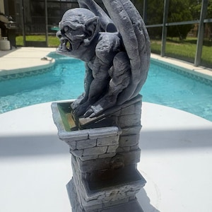 Puede incluir: Una estatua de g&aacute;rgola gris posada sobre un pedestal con textura de piedra y un peque&ntilde;o cuenco. La g&aacute;rgola tiene alas grandes, dientes afilados y una superficie texturizada. El pedestal presenta un patr&oacute;n de ladrillo y un anillo.