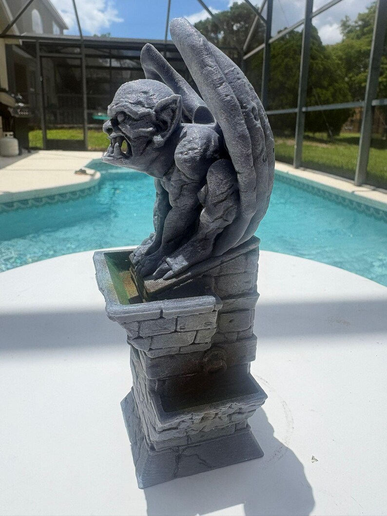 Puede incluir: Una estatua de g&aacute;rgola gris posada sobre un pedestal con textura de piedra y un peque&ntilde;o cuenco. La g&aacute;rgola tiene alas grandes, dientes afilados y una superficie texturizada. El pedestal presenta un patr&oacute;n de ladrillo y un anillo.