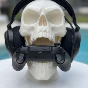 May include: A white human skull model wearing black over-ear gaming headphones. The skull is holding a black Xbox video game controller in its open mouth. The controller features a visible charging port and a small circular sync button on the top.