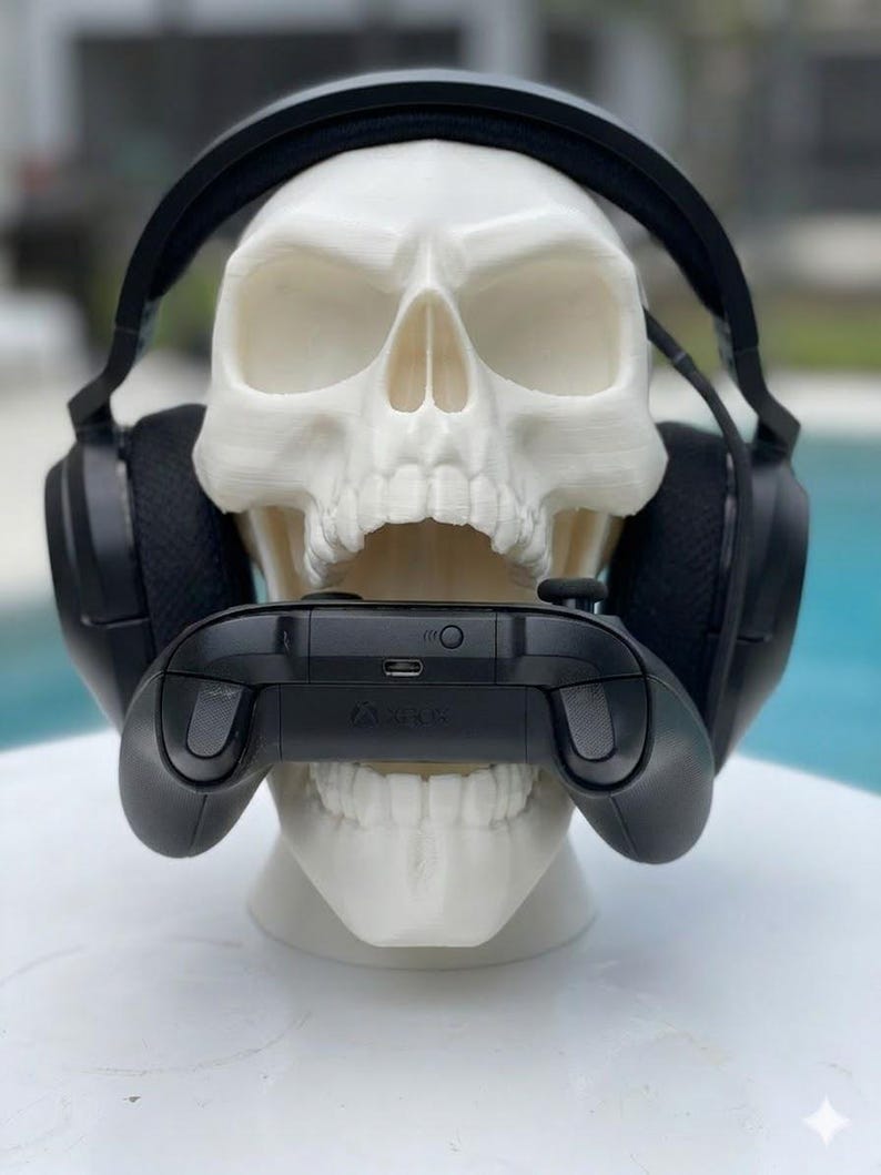 May include: A white human skull model wearing black over-ear gaming headphones. The skull is holding a black Xbox video game controller in its open mouth. The controller features a visible charging port and a small circular sync button on the top.