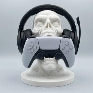 May include: A white skull-shaped stand holds a pair of black over-ear gaming headphones with a microphone boom and a white and black wireless video game controller. The skull rests on a circular base, providing a display for gaming accessories.