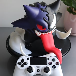 May include: A hybrid creature figurine featuring a purple and black split design with a long red tongue, sharp white teeth, and a red eye, positioned behind a white video game controller with black buttons and a central PlayStation logo.