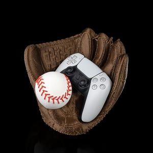Puede incluir: Un guante de b&eacute;isbol de cuero marr&oacute;n que sostiene una pelota de b&eacute;isbol blanca con costuras rojas y un mando de videojuego inal&aacute;mbrico blanco y negro con el logotipo de PlayStation y s&iacute;mbolos geom&eacute;tricos ic&oacute;nicos sobre fondo negro.
