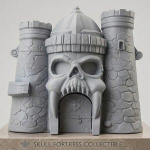 Op de afbeelding: Een grijze 3D-geprinte schedelfort-collectible met twee torens met steentextuur die een centrale schedelstructuur met een poort flankeren. De deur toont een schedel- en kruisbeenderenembleem. De basis is gelabeld met SKULL FORTRESS COLLECTIBLE.