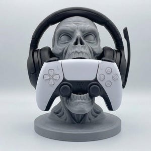 May include: A gray skull-shaped stand holding a black gaming headset over its cranium and a white and black wireless game controller in its open mouth. The skull rests on a circular base, providing a structured display for gaming accessories.
