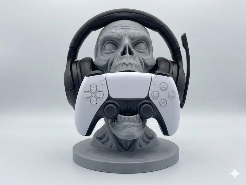 May include: A gray skull-shaped stand holding a black gaming headset over its cranium and a white and black wireless game controller in its open mouth. The skull rests on a circular base, providing a structured display for gaming accessories.