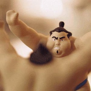 May include: A tan-colored figurine of a sumo wrestler with a topknot hairstyle, intense facial expression, and raised arms. The figure features a dark brown textured element near the neck and a thin blue band visible at the lower edge of the torso.